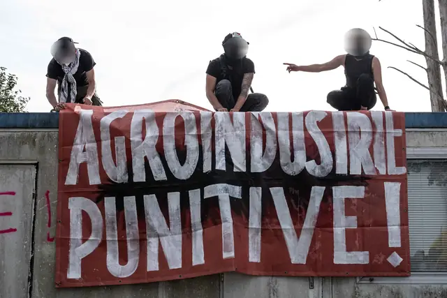 ثلاثة أشخاص ملثّمين على سطح فوق لافتة حمراء «الصناعة الزراعية العقابية!» (Agro-industrie punitive !)