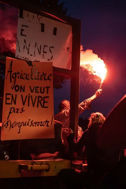 De noche, una bengala roja ilumina el cartel «Agricultura: ¡queremos vivir de ella, no envenenarnos!» («Agriculture : on veut en vivre, pas s’empoisonner !»)