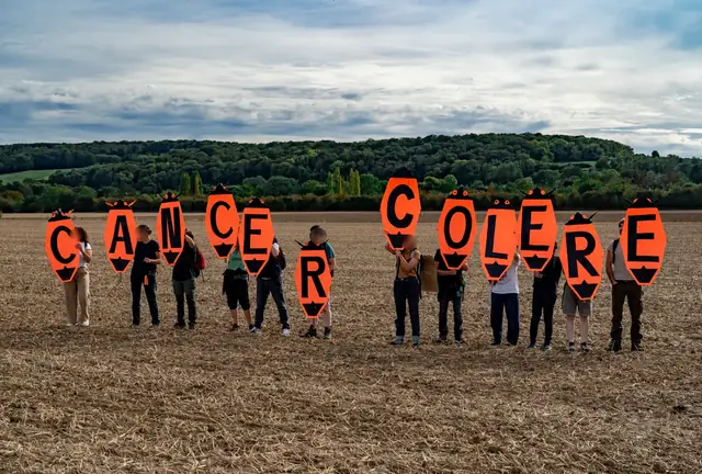 Manifestantes en fila en un campo forman las palabras «Cáncer rabia» («Cancer colère») con grandes letras naranjas