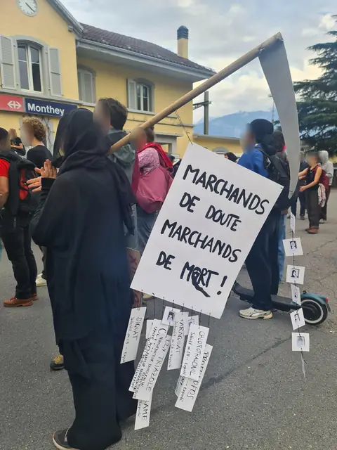 شخص متنكر في هيئة ملاك الموت يحمل لافتة «تجّار الشك، تجّار الموت!» («Marchands de doute, marchands de mort !») أمام المحطة
